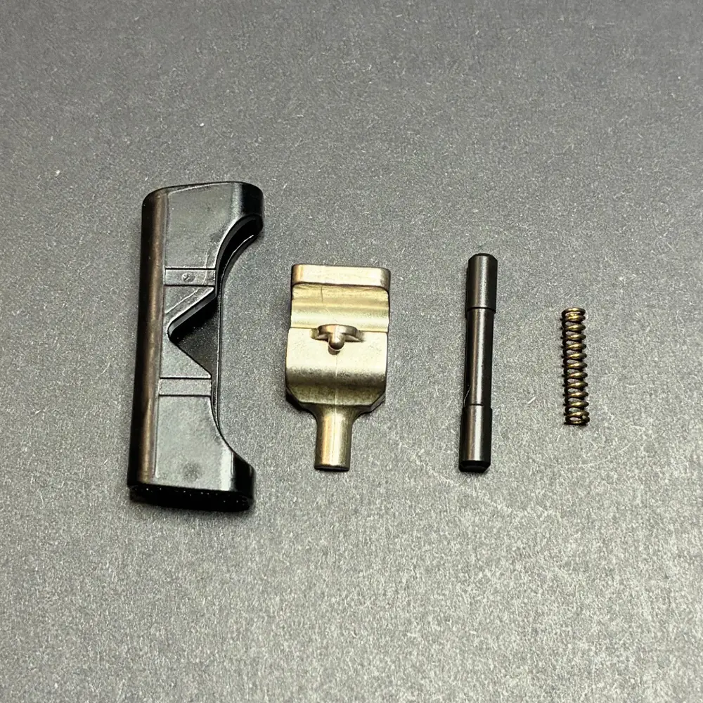 Close-up of a pistol magazine release kit with the magazine catch, release button, retaining pin, and spring arranged in sequence on a gray background.