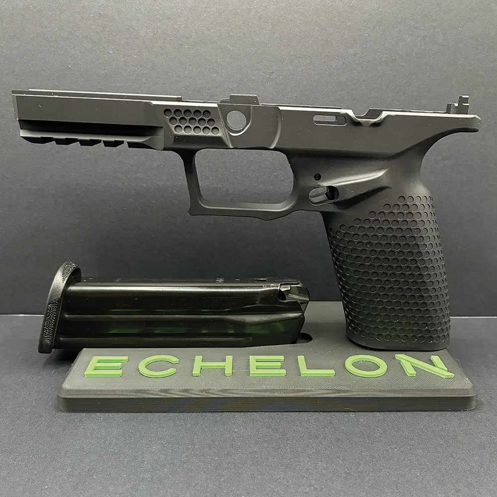 Side view of a black precision-machined pistol frame with a textured grip and vented front section, displayed on an 'ECHELON' stand beside a detached magazine against a black background.