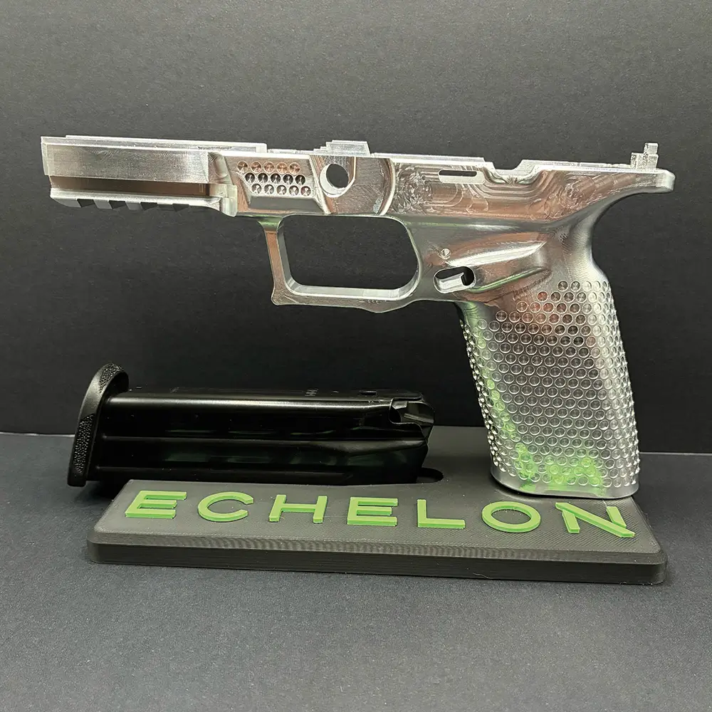 Close-up of a precision-machined metal pistol frame with textured grip and engraved details, displayed on a black stand labeled 'ECHELON' beside a detached magazine against a black background.