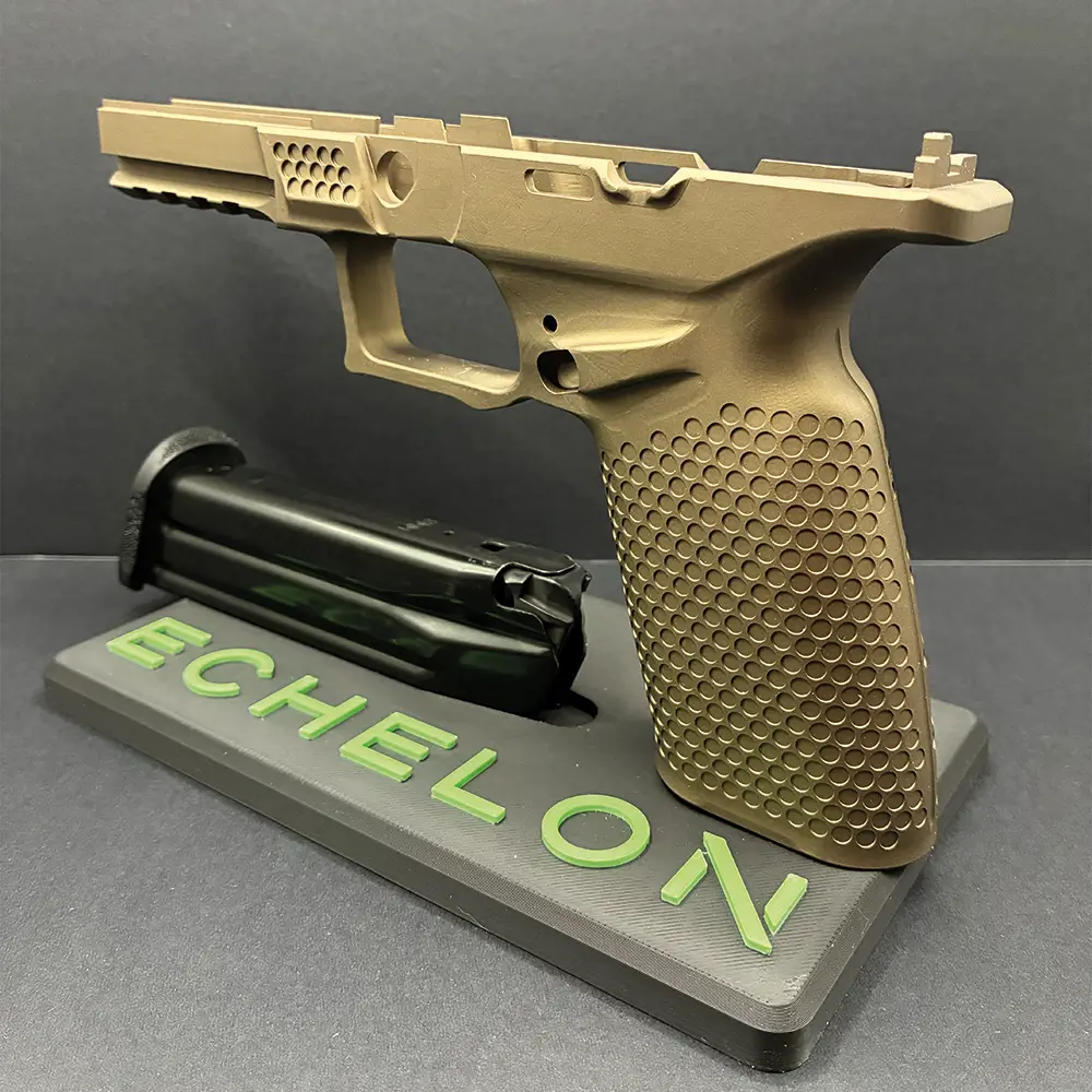 Angled view of a tan-coated precision-machined pistol frame with a textured grip and vented front section, displayed on an 'ECHELON' stand beside a detached magazine against a black background.