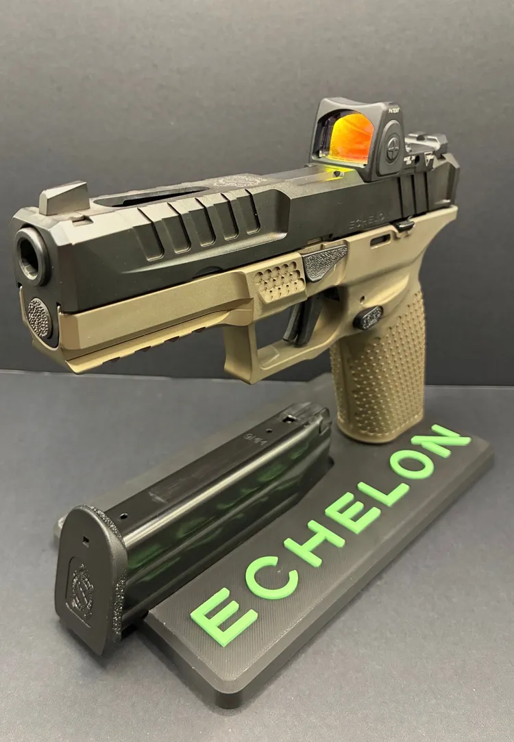 An angled, front three-quarter view of a complete Springfield Armory Echelon pistol featuring a black slide and a tan textured grip module, equipped with a red dot optic showing a bright orange reflection on the lens. The firearm is mounted upright on a dark grey display stand with a black extended pistol magazine resting flat on the base beneath the frame. The front of the display stand features the word "ECHELON" in raised, bright green lettering, all set against a dark background.