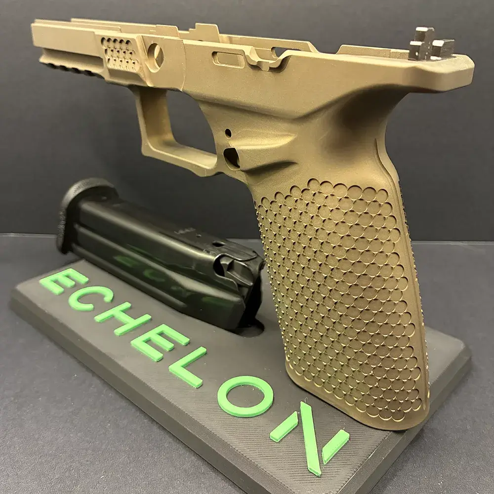 A tan Springfield Armory Echelon pistol grip module, missing the slide and internal components, mounted upright on a grey display stand. The stand features the word "ECHELON" in raised, bright green lettering. A black firearm magazine lies flat on the stand beside the grip module, set against a dark background.