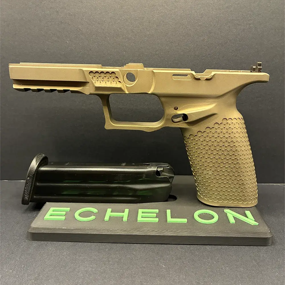 A side-profile view of a stripped, tan Springfield Armory Echelon pistol grip module, missing its slide and internal components, displayed upright on a dark grey stand. A black extended pistol magazine rests flat on the base of the stand beneath the frame. The front of the display stand features the word "ECHELON" in raised, bright green lettering, all set against a dark grey background.