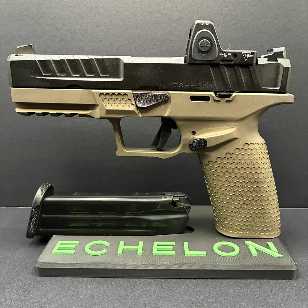 A side-profile view of a complete Springfield Armory Echelon pistol featuring a black slide and a tan textured grip module, equipped with a Trijicon red dot optic. The firearm is mounted upright on a dark grey display stand with a black extended pistol magazine resting flat on the base beneath the frame. The front of the display stand features the word "ECHELON" in raised, bright green lettering, all set against a dark background.